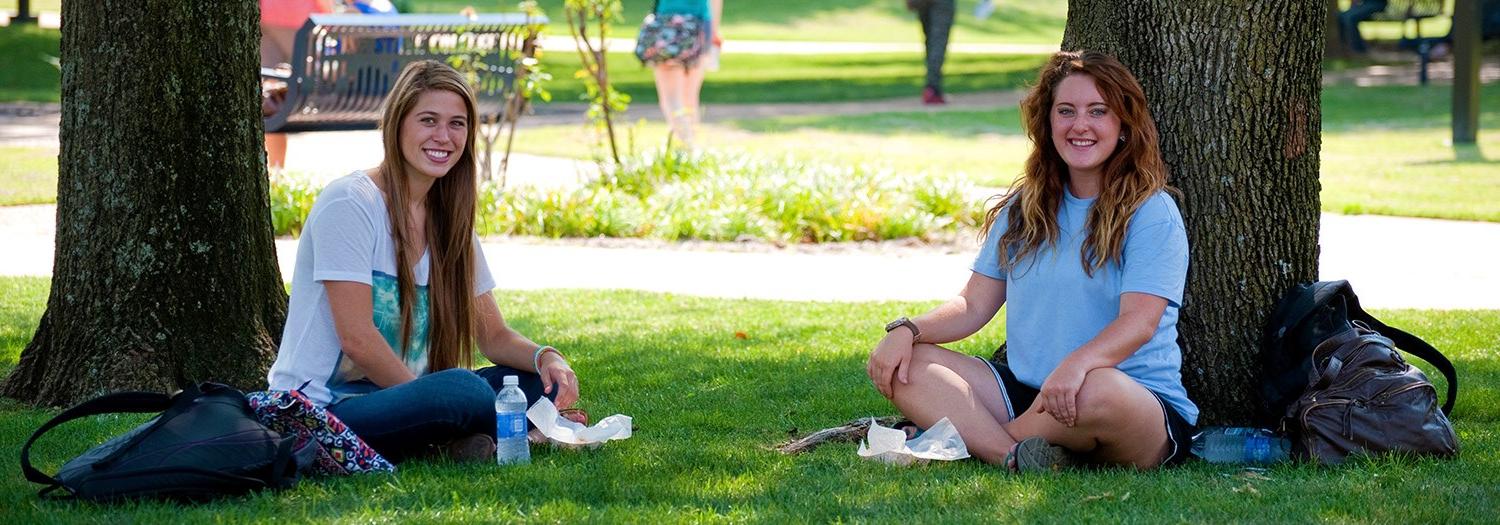 Two students seated in the shade of large trees in the central mall area of the campus